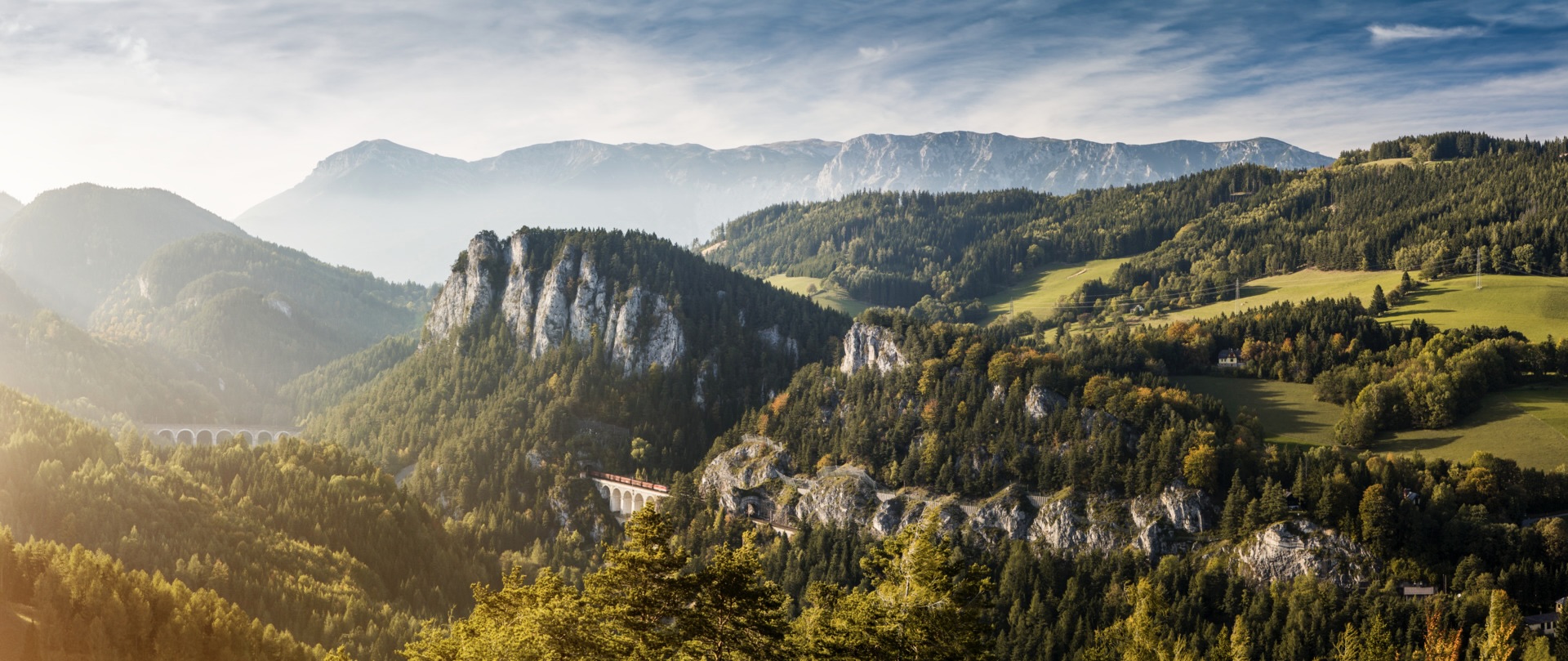 Panorama der Region Reichenau-Rax: Zug auf dem Kalte-Rinne-Viadukt der Semmeringbahn vor bewaldeten Hängen und den Kalkwänden des Rax-Schneeberg-Massivs im Morgenlicht